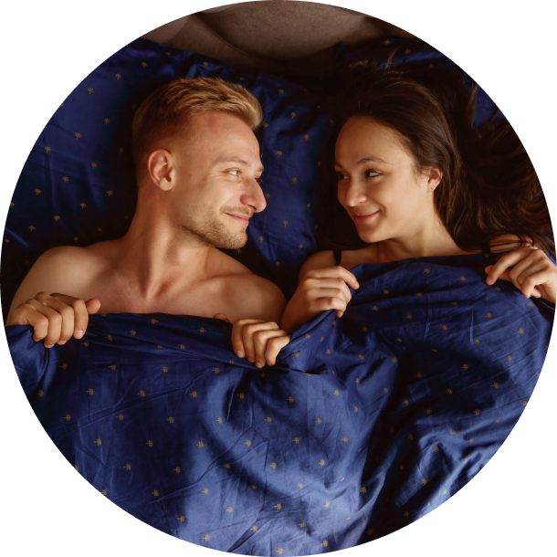 married couple lying in bed under sheets, sharing a playful, cheeky, secretive smile after intimacy